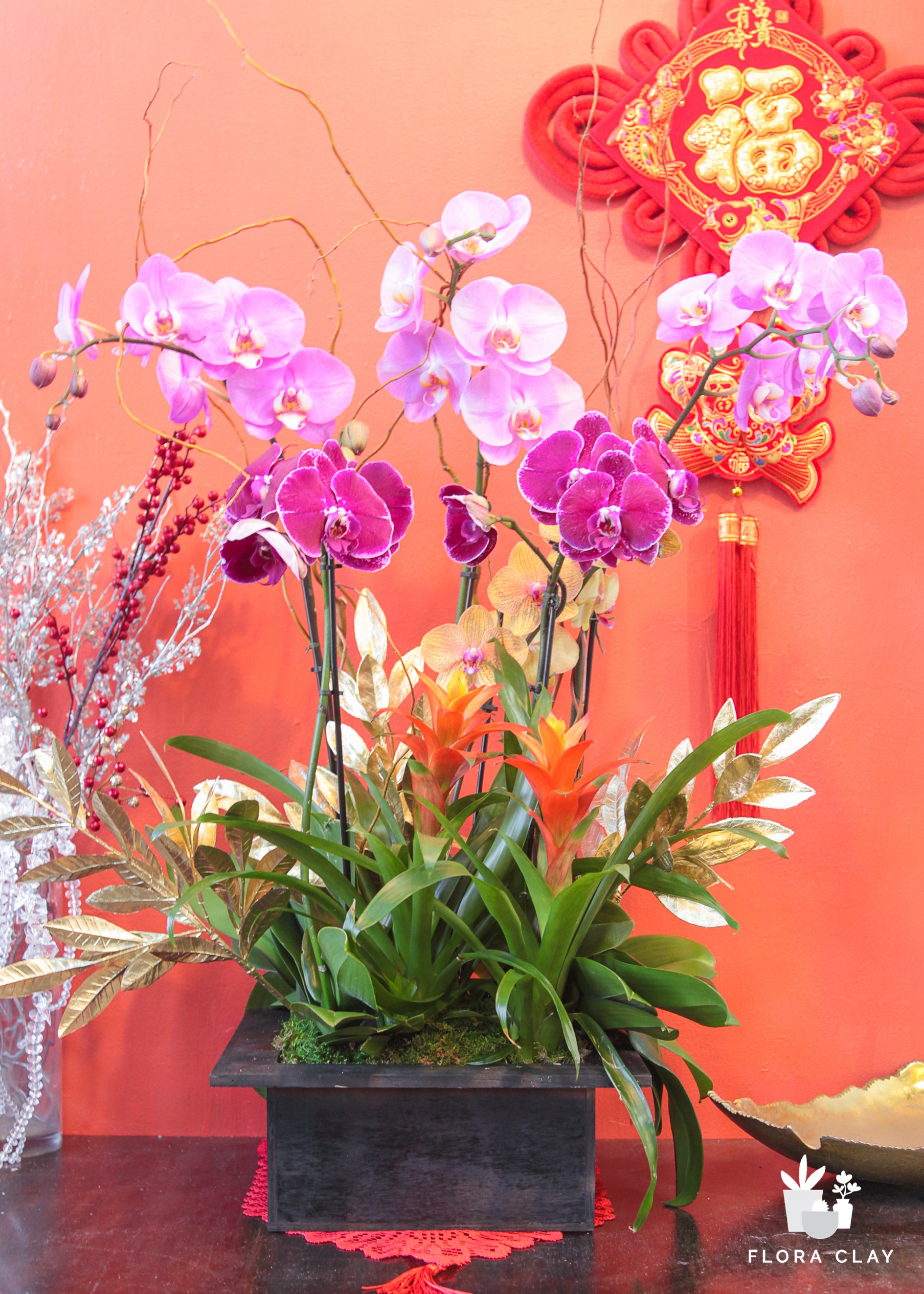 A floral arrangement featuring pink orchids and other greenery, presented in a square container, with decorative elements and a 'Happy Lunar Year' sign in the background.