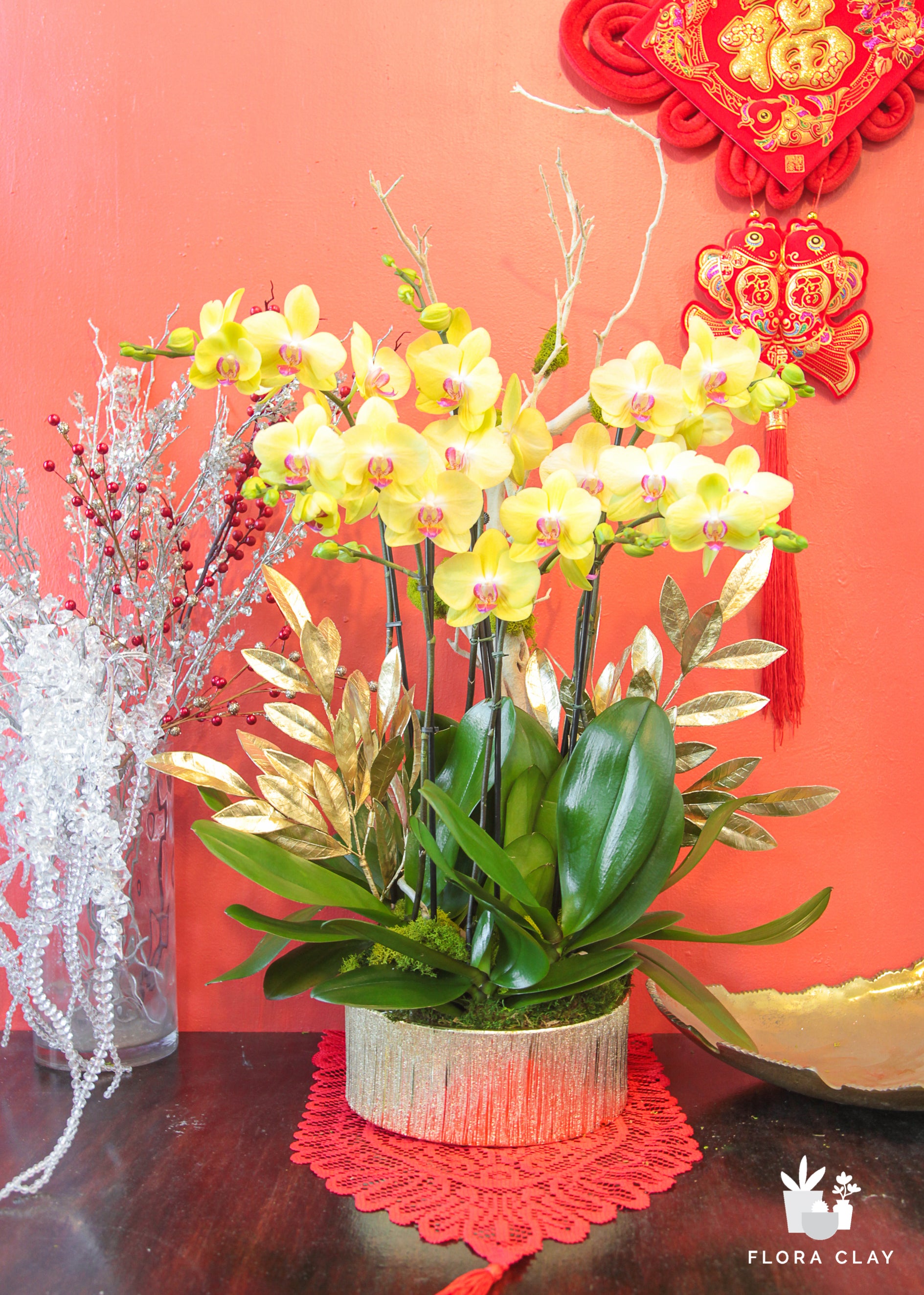 A floral arrangement featuring yellow and pink orchids with green leaves, displayed in a red vase, against a red background with a Chinese New Year decoration in the background.