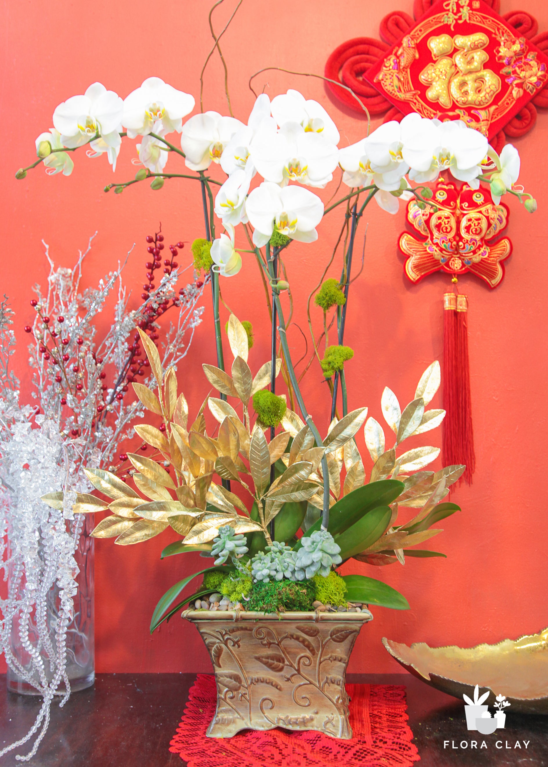 An orchid arrangement featuring a combination of pink and white orchids with green leaves, gold-colored foliage, and decorative elements in a traditional Chinese vase, displayed against a red and gold background with Chinese New Year decorations.