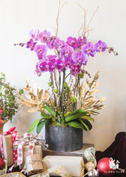 A artificial floral arrangement featuring pink orchids, gold faux leaves, and berries, displayed in a black container.