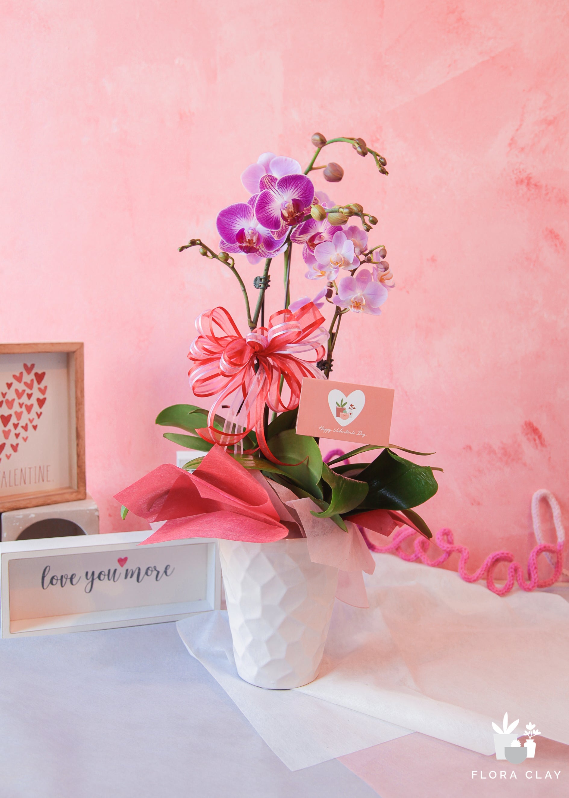 A floral arrangement with white and purple orchids and green foliage, wrapped in a pink ribbon bow, with a pink and green message card, presented in a white vase.