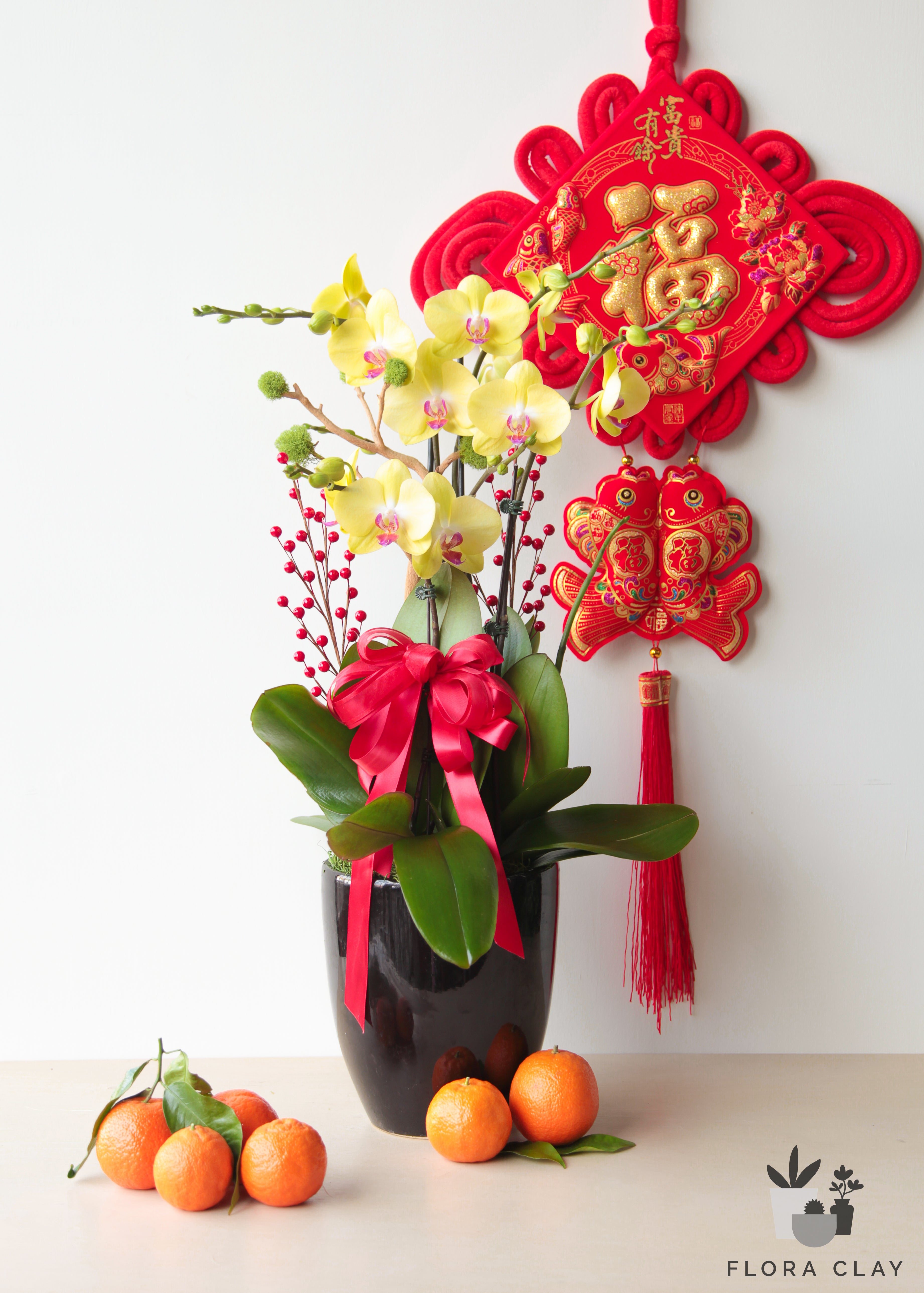 An orchid arrangement with red and gold flowers, featuring a Chinese New Year theme with a decorative backdrop and bamboo accents, displayed with citrus fruits and a hanging lantern.