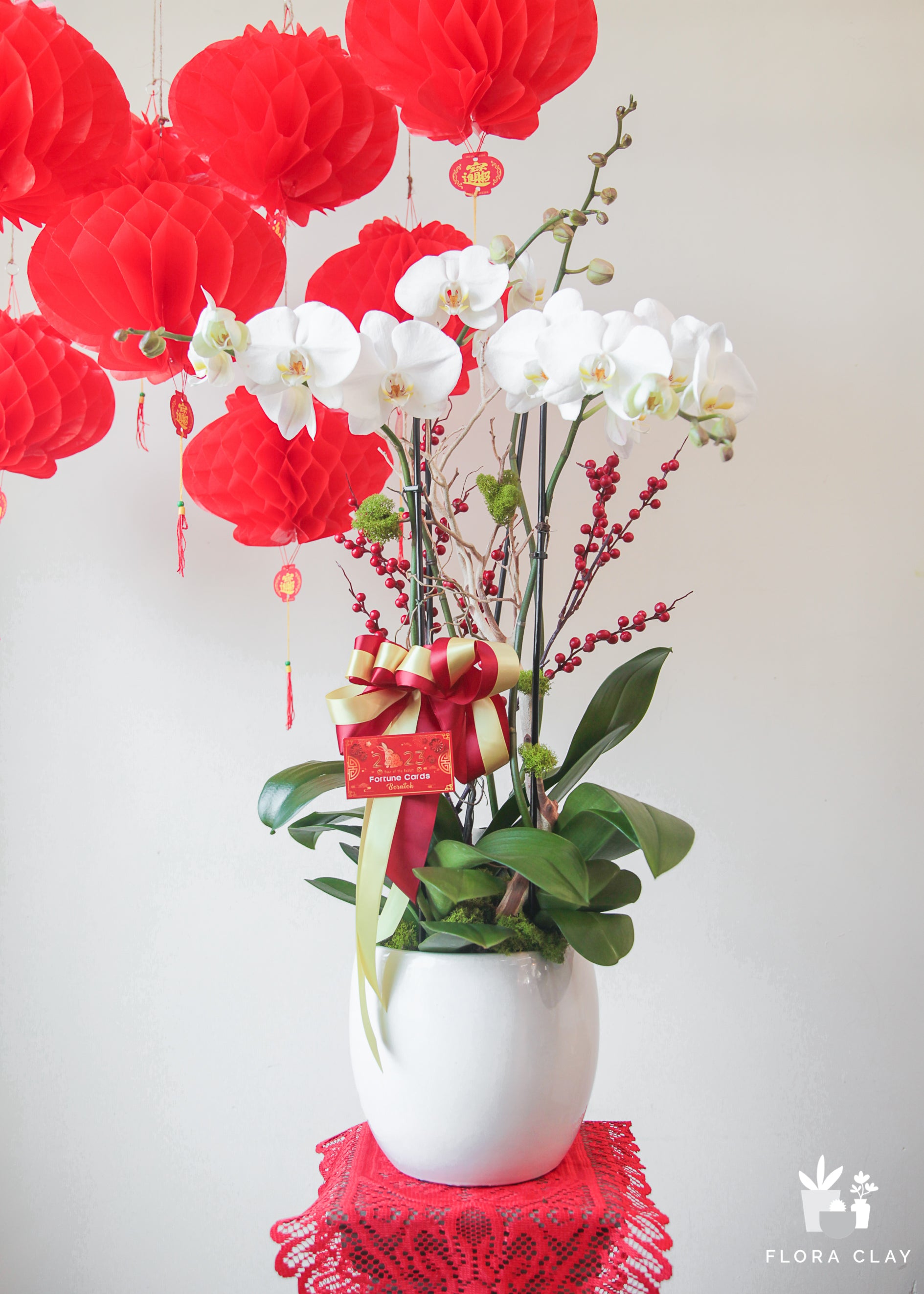 A decorative floral arrangement featuring white and red flowers, with a prominent orchid, tulips, and other blooms, presented in a white pot on a red lace placemat against a white wall.
