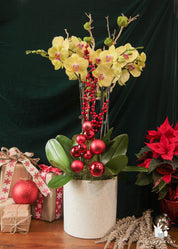 A floral arrangement featuring yellow orchids, red poinsettias, and green leaves in a white vase, with Christmas decorations in the background.