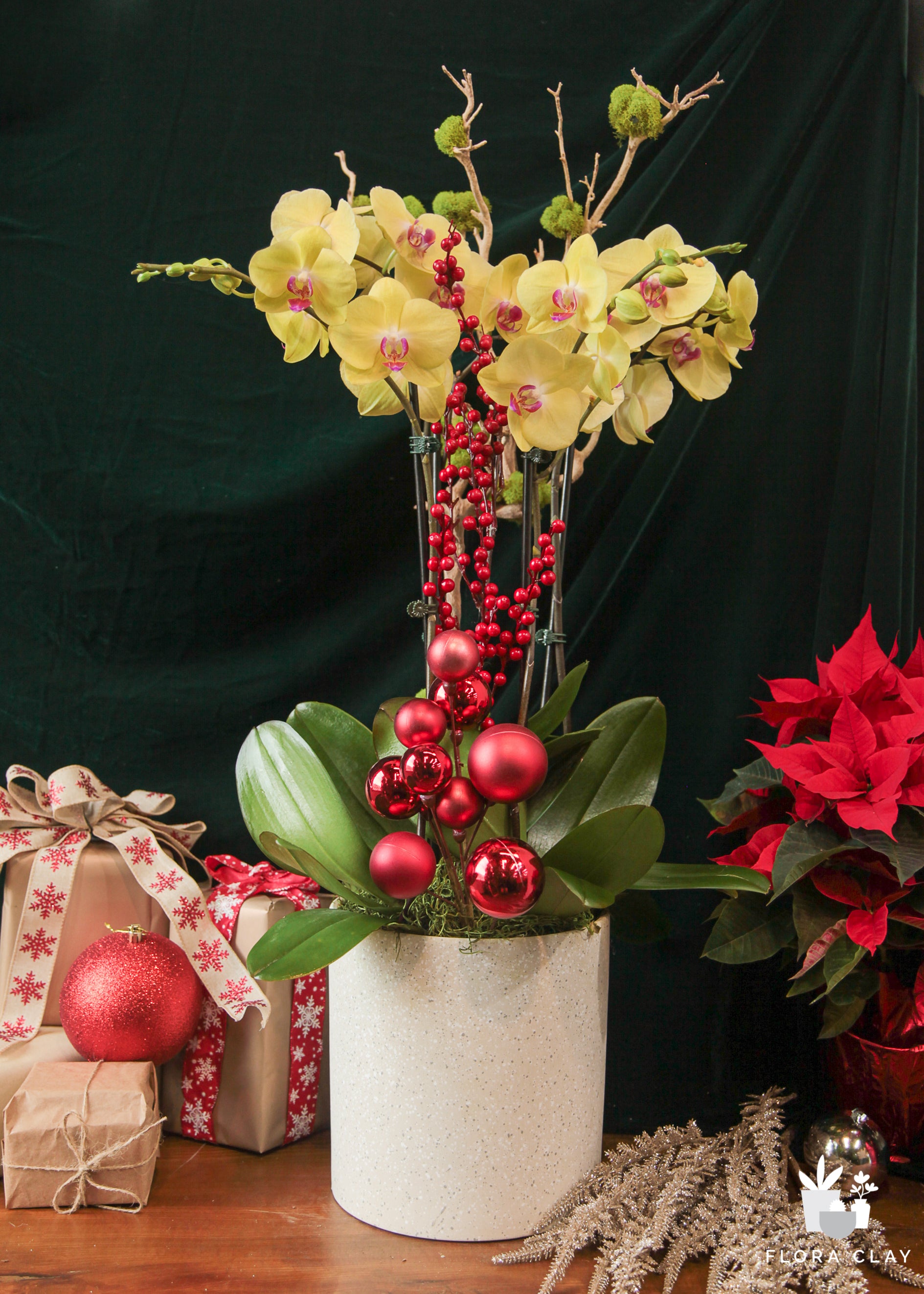 A floral arrangement featuring yellow orchids, red poinsettias, and green leaves in a white vase, with Christmas decorations in the background.