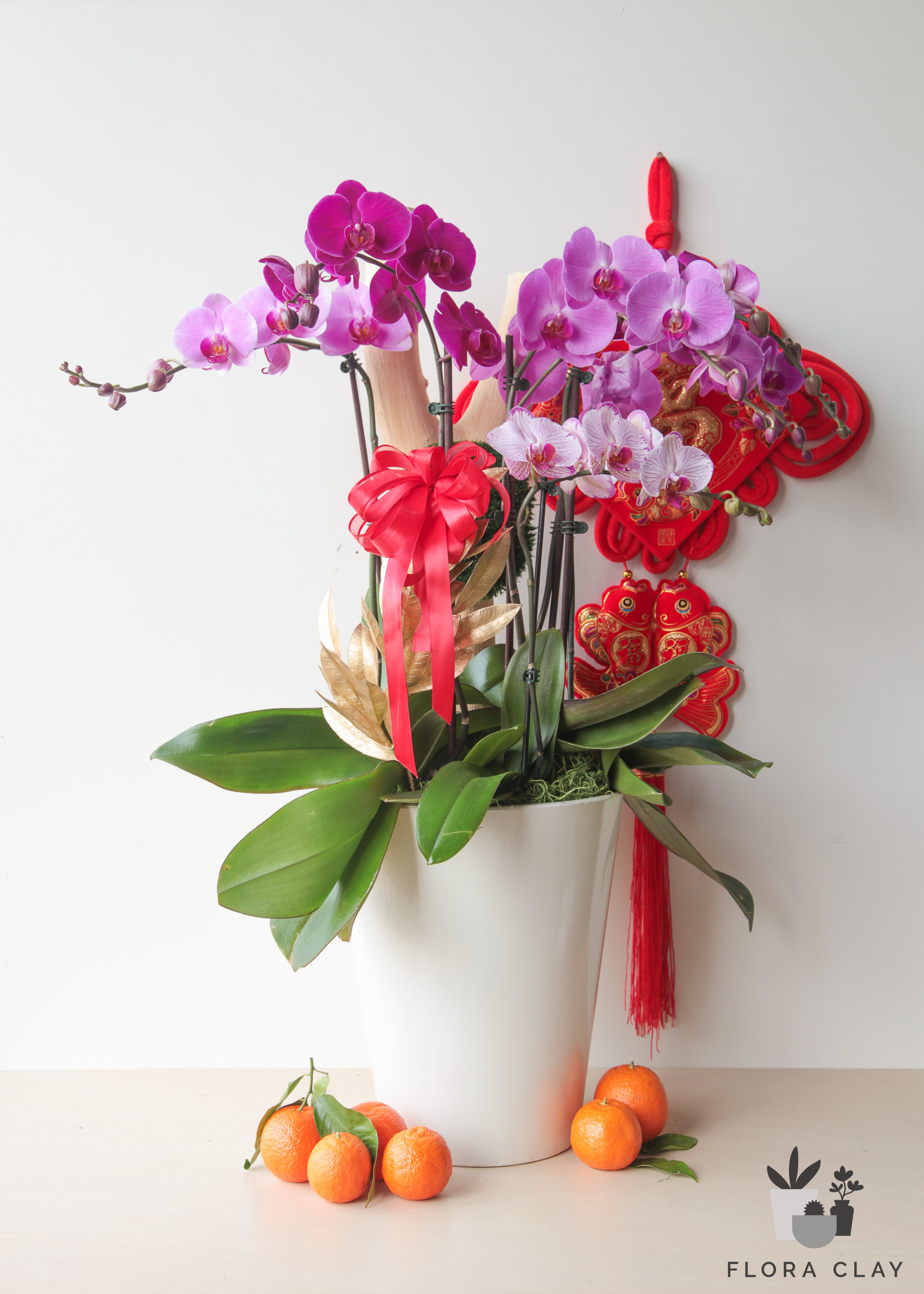 An orchid arrangement with pink and red flowers, green leaves, and decorative elements, presented in a white vase, with a small bow and orange fruits beside it.