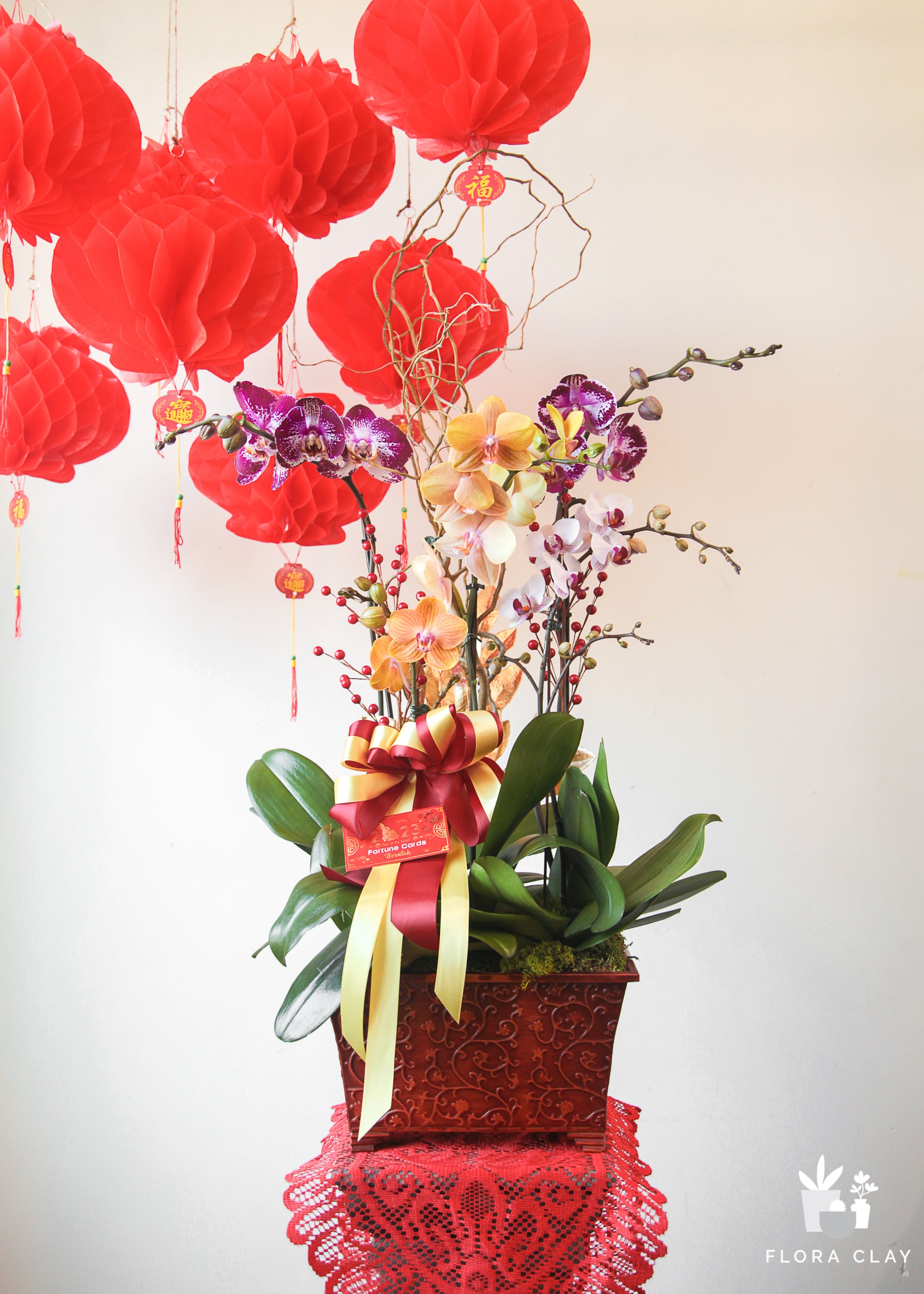 An orchid arrangement with pink and red flowers, featuring a bunch of pink orchids and decorative leaves, presented in a red container with a floral lace cover.