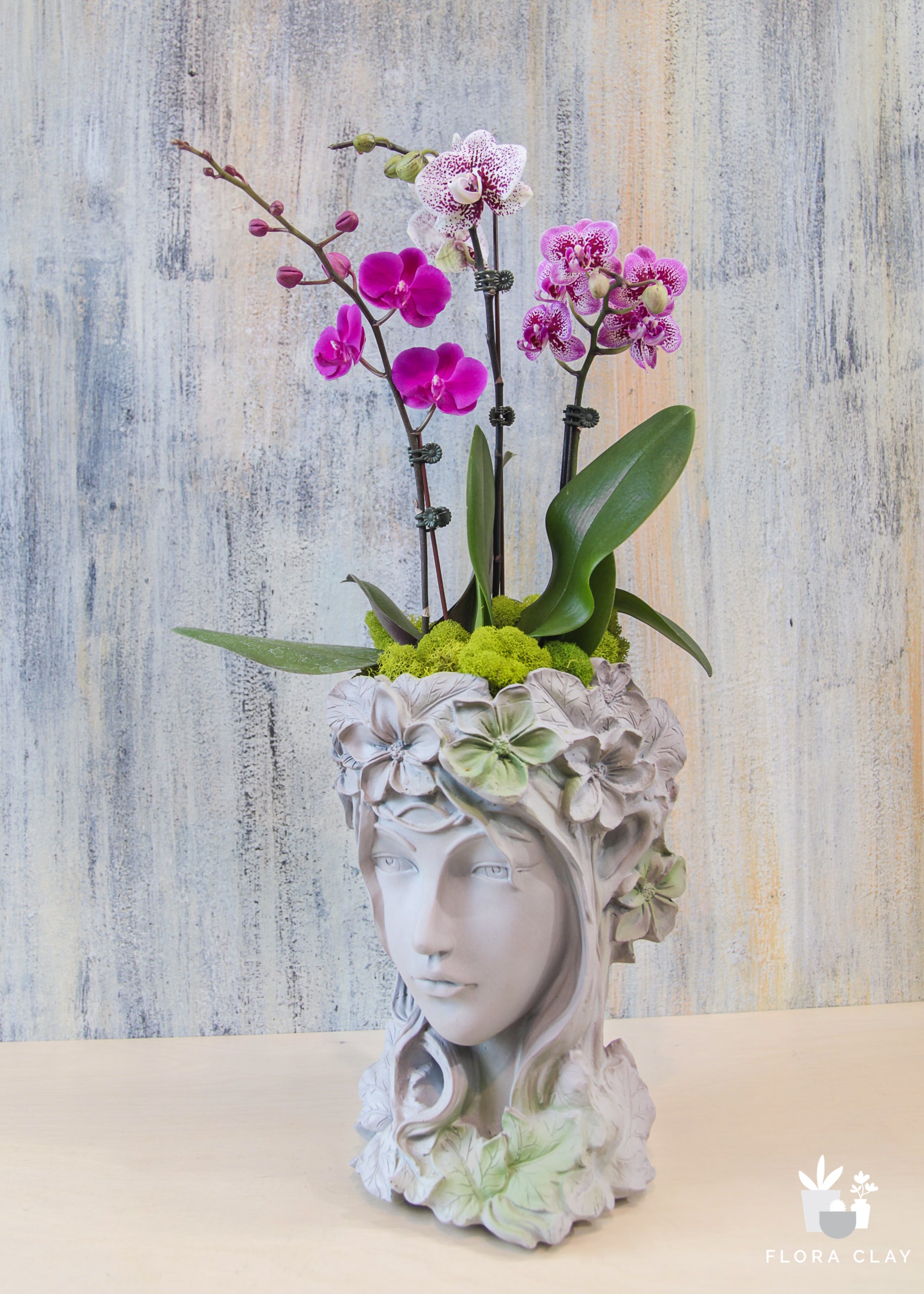 A decorative vase in the shape of a woman's head, with a floral design, containing a pink orchid arrangement.